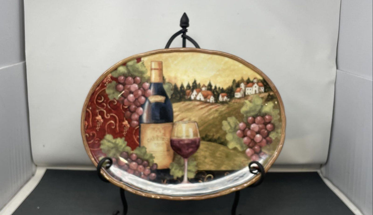 Oval decorative platter painted with grapes, wine bottle, filled glass and village, on a black metal stand, view 2